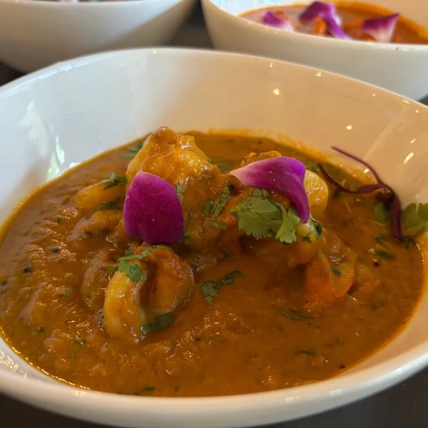 Indian curry with shrimp and cilantro, garnished with purple edible flowers in a white bowl.